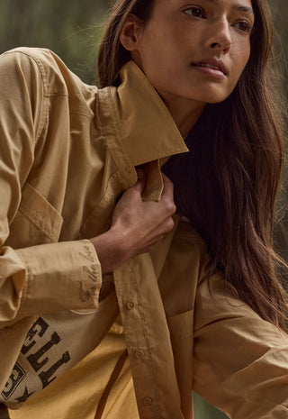 A close-up lifestyle shot of a model wearing a tan utility shirt jacket with a high collar and a matching tan top underneath. The jacket features a chest pocket and a sleeve with a black graphic print.