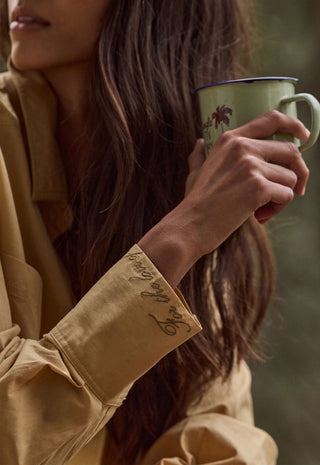 A close-up lifestyle shot of a woman holding a light green enamel mug with a dark rim and palm tree print, wearing a tan button-up shirt with embroidered script on the cuff.