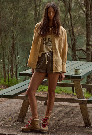 A woman stands outdoors at a wooden picnic table in a wooded setting, wearing a tan overshirt, a graphic T-shirt, brown shorts, and tan lace-up boots.
