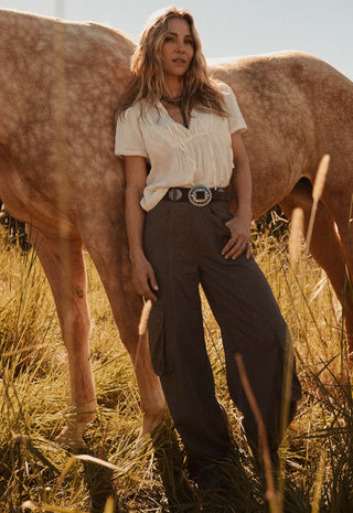 A woman stands in a field of dry grass next to a horse, wearing a cream short-sleeve blouse, dark brown wide-leg cargo pants, and a silver belt.