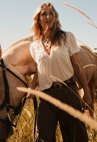  wearing a cream short-sleeve blouse tucked into dark brown wide-leg trousers with a western-style belt and large buckle.