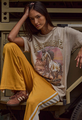 A woman models a beige graphic t-shirt with a western horse design and the text 'FOR THE LOVE OF THE WILD' paired with mustard yellow pants and brown sneakers, posing on a rustic wooden bench.