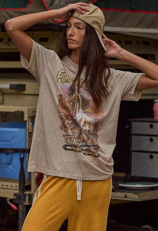 A woman wearing a beige graphic t-shirt with a cowboy on the front, paired with mustard yellow sweatpants and a khaki cap, posing in front of a rustic vehicle.