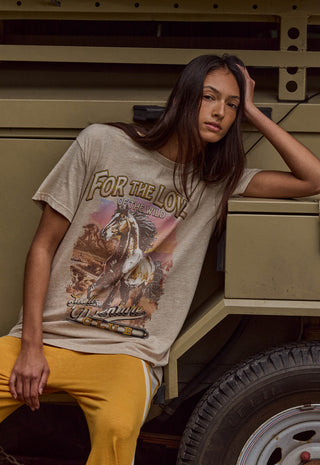 A woman wearing a beige graphic t-shirt with a western theme and yellow pants, leaning against a military-style vehicle.