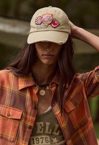 a-woman-wearing-a-beige-baseball-cap-with-multiple-embroidered-patches-an-orange-and-brown-plaid-flannel-shirt-and-a-graphic-tank-top-posed-outdoors-against-a-blurred-green-background