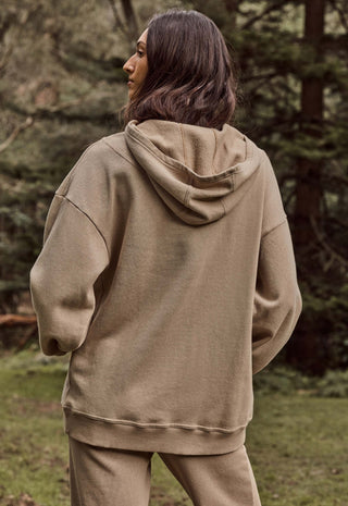 A woman wearing an oversized taupe-brown hooded sweatshirt and matching sweatpants, photographed from behind in an outdoor forest setting.