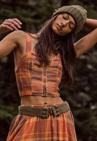 a-woman-wearing-a-rust-orange-and-brown-striped-knit-top-with-a-button-front-a-matching-plaid-skirt-and-an-olive-green-beanie-posed-outdoors-against-a-dark-blurred-forest-background