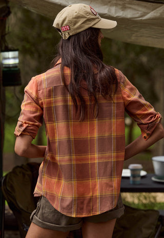 a-woman-viewed-from-behind-wearing-a-tan-baseball-cap-a-rust-orange-and-brown-plaid-flannel-shirt-and-olive-shorts-in-an-outdoor-camping-setting-under-a-canopy