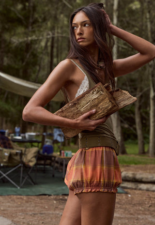 a-woman-standing-outdoors-in-a-wooded-campsite-setting-holding-a-piece-of-wood-she-is-wearing-a-sleeveless-lace-trim-top-an-olive-green-belt-and-a-rust-orange-plaid-ruffled-skirt