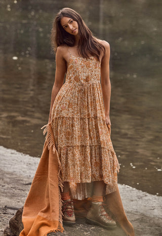A woman stands outdoors by a body of water wearing a sleeveless, tiered maxi dress with an allover floral pattern in warm tan, rust, and cream tones. The dress has thin straps and a fringed hem, and she is also wearing an orange-brown fringed throw blanket draped over one side.