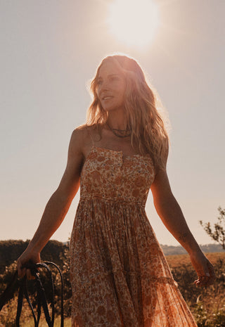 A woman outdoors in warm golden-hour sunlight wearing a strapless floral maxi dress, holding a dark woven strap in one hand. The background is a bright sunlit sky with blurred brush and hills.