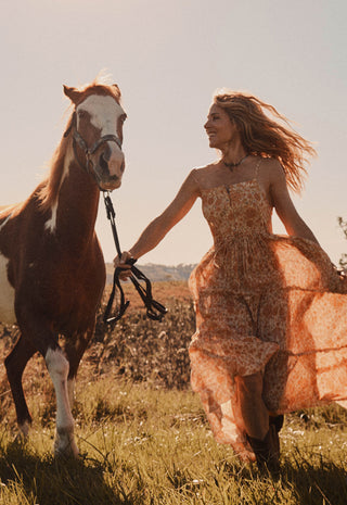 A woman in a flowing peach floral dress walks beside a brown and white horse in an open field, holding the horse's reins under warm golden sunlight.