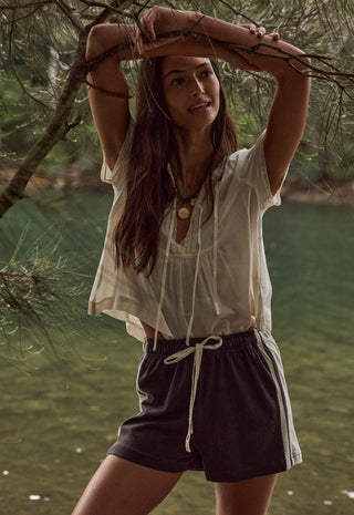 A woman outdoors by a body of water under tree branches, wearing a loose off-white short-sleeve blouse and dark charcoal drawstring shorts with white side stripes.