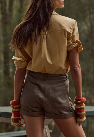 a-woman-shown-from-the-back-wearing-a-tan-short-sleeve-button-up-shirt-and-brown-shorts-with-striped-knit-fingerless-gloves-standing-outdoors-against-a-blurred-forest-background